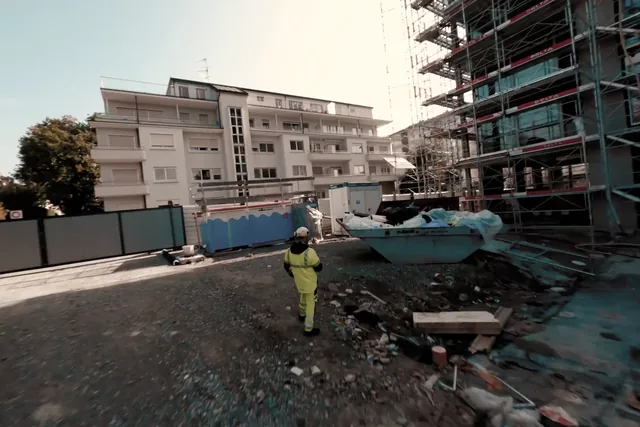 On a construction site, a worker in a yellow vest walks towards a full M. Ducret SA skip in front of a building under renovation, still from the Nereva film
