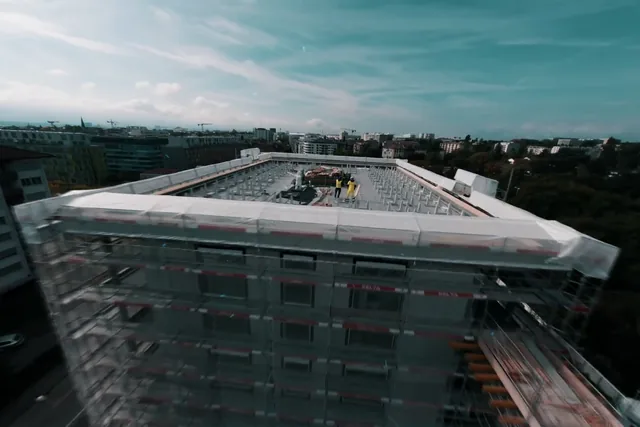 Aerial view of a building under construction wrapped in white sheeting, two workers on the roof and the cranes of Geneva on the horizon, still from the Nereva film